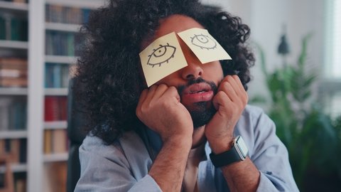 Lazy office worker napping at workplace covering eyes with sticky notes, snoring funny moving lips. Inefficient tired male employee pretends working sleeping with stickers on face sits at desk.