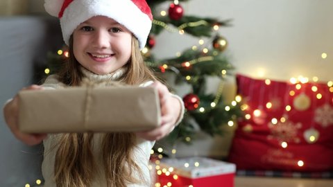 Cute little girl smiling giving handmade stylish simple Christmas gift box in craft paper at home near Christmas tree. Happy child girl with present. Winter holidays. DIY, zero waste concept