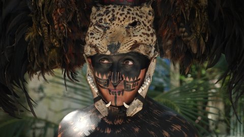 Tulum, Mexico - Nov 20 2021: Traditional indian aztec shaman with skull and jaguar skin with makeup wearing a plume headdress with real animal skulls, playing conch shell trumpet, with maguey plants.
