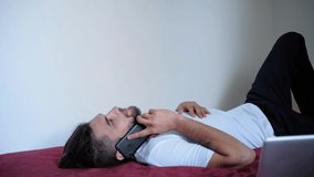 Talk on the phone and discuss important work things. Caucasian male freelancer with beard and dreadlocks works online at home. Lying on red blanket on bed looking up and smiling. 4K footage.