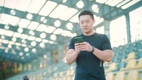 happy successful Asian male athlete scrolling using typing mobile phone, browses makes purchases online use smartphone smiling, outside at the sports stadium. athletic man read message text Copy space
