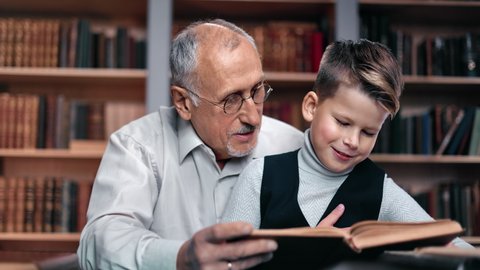 Closeup happy family reading interesting book at home public vintage library spending time together positive emotion. Smiling 70s grandfather and cheerful grandchild doing homework literature textbook
