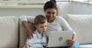 Adorable little boy sit on sofa at living room in warm emb s of caring Indian mom play education online game in laptop web app. Loving young mum hug small son browsing social network on pc device