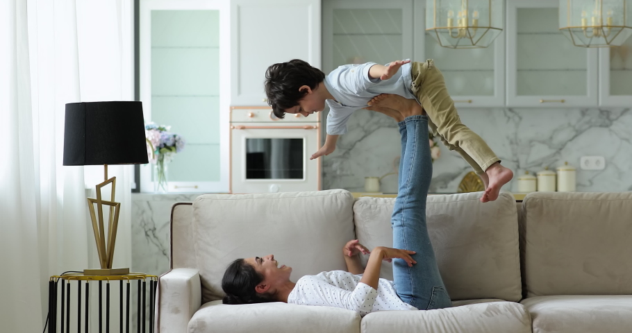 Mommy I can fly. Happy Indian mother older sister lying on back on cozy sofa having fun with little kid lifting excited child boy in air on outstretched legs. Active mom play airplane with small son Royalty-Free Stock Footage #1082693284