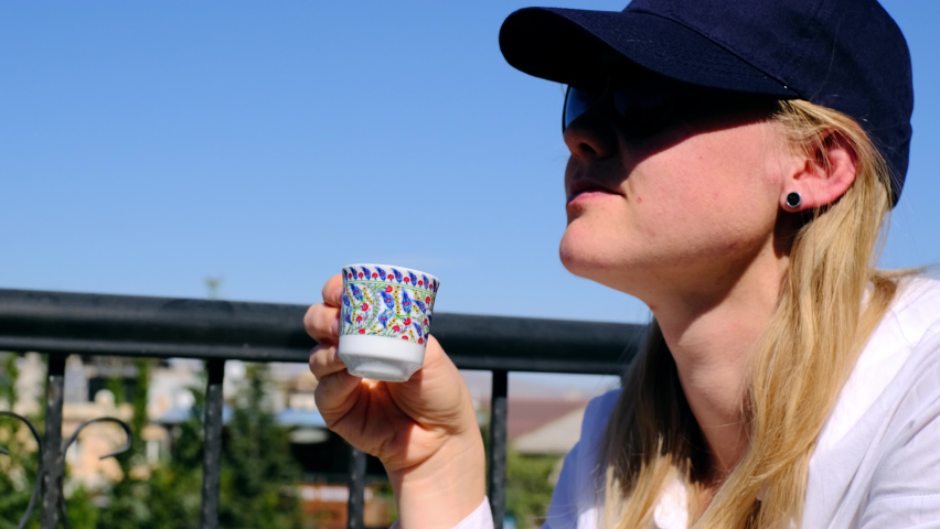 young woman in white shirt and blue cap drinks Turkish coffee from traditional cup and washed it down with water. lady sits on terrace on hot day and quenches her thirst with black coffee from cup.