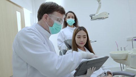 Caucasian male and femal dentist with face mask use tablet to advice how to fix teeth to smiling asian woman patient at modern dental clinic. Dentistry business during covid 19 pandemic.
