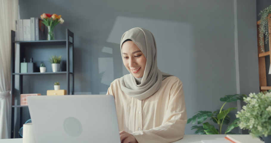 Attractive cheerful young Asian muslim businesswoman in hijab with casual sitting at the desk use laptop remote working in living room at home. People religious Islam lifestyle concept. Royalty-Free Stock Footage #1082707204