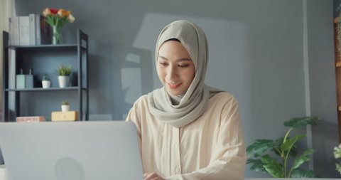 Attractive cheerful young Asian muslim businesswoman in hijab with casual sitting at the desk use laptop remote working in living room at home. People religious Islam lifestyle concept.