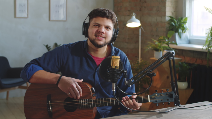 Young cheerful Hispanic man in headphones sitting with guitar in recording studio, smiling, waving at camera and speaking into microphone while giving online music class