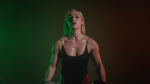 A fitness model in a bodysuit is looking at the camera. She is doing a shoulder exercise. She is holding dumbbells and spreading them out to the sides. An orange and green light is shining on her. 4K