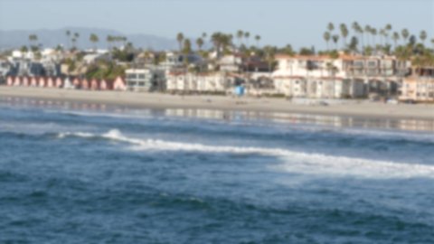 Defocused pacific ocean coast from pier. Sea water waves tide, shore sand. Beachfront vacations resort. Waterfront Oceanside, California USA. Tropical palms, cottages on beach. Reflection in littoral.