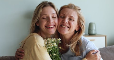 Portrait of happy aged mom stand with closed eyes hold smiling young daughter in tender embraces receiving bunch of flowers from beloved adult child. Affectionate mum grownup kid cuddle look at camera