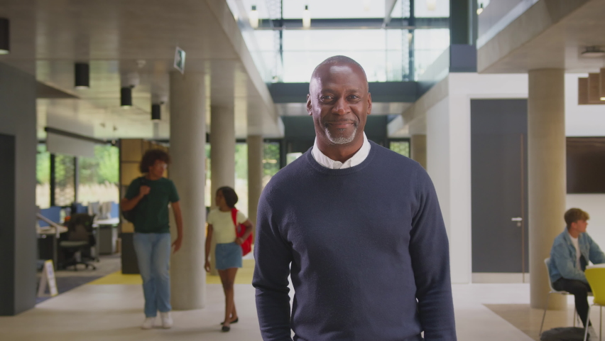 Portrait of smiling mature male tutor standing in busy university or college building with students- shot in slow motion