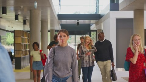 Male and female tutors walking along busy college corridor looking at digital tablet surrounded by students - shot in slow motion