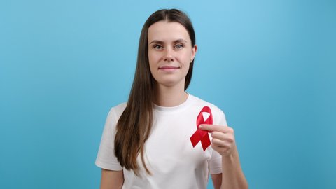 Concept World AIDS and hiv day. Young brunette woman holds red ribbon, looking at camera, isolated on blue color background studio. Blood transfusion and donation. Hemophilia and World Health concept