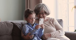 Loving old grandma sit on couch hug little grandkid watch girl playing phone game. Two diverse age female generations of one family senior granny small granddaughter discuss funny video on web channel