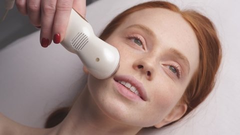 an attractive woman in a cosmetology clinic receives a facial skin phonophoresis procedure. The cosmetologist moves the manipulator across the face.