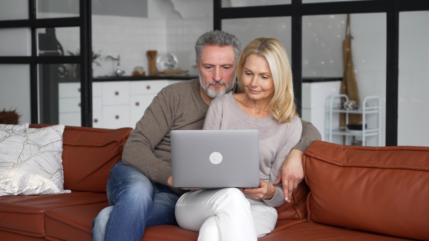 Middle-aged couple in love using laptop sitting on the couch at home. Cheerful senior couple watching movie together, smiling and hugging at cozy living room, web surfing and spends time online Royalty-Free Stock Footage #1082733151