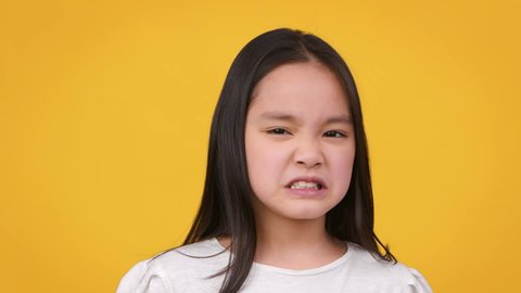 Eww. Close up portrait of cute little asian girl frowning her face, expressing disgust and aversion, smelling or tasting something awful, orange studio background, slow motion