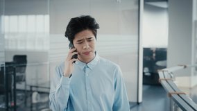 Sad Asian Business Guy Talking On Phone Walking In Modern Office Indoors. Mobile Communication, People And Gadgets Concept. Tracking Shot, Slow Motion