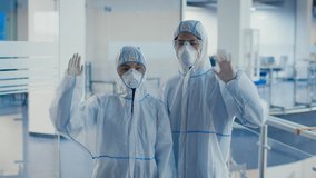 Asian Couple Of Laboratory Workers Wearing Protective Hazmat Suits And Respirator Face Masks Waving Hello To Camera Standing In Modern Hospital Or Scientific Center Indoors. Slow Motion