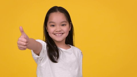 Kids approval. Studio portrait of cute positive little asian girl gesturing thumb up and sincerely smiling to camera, posing over orange background with free space, slow motion