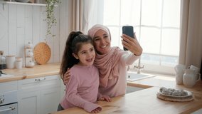 Selfie With Mom. Joyful Middle Eastern Mother In Hijab And Little Daughter Using Smartphone Taking Photos Of Themselves Posing Having Fun Together In Kitchen At Home