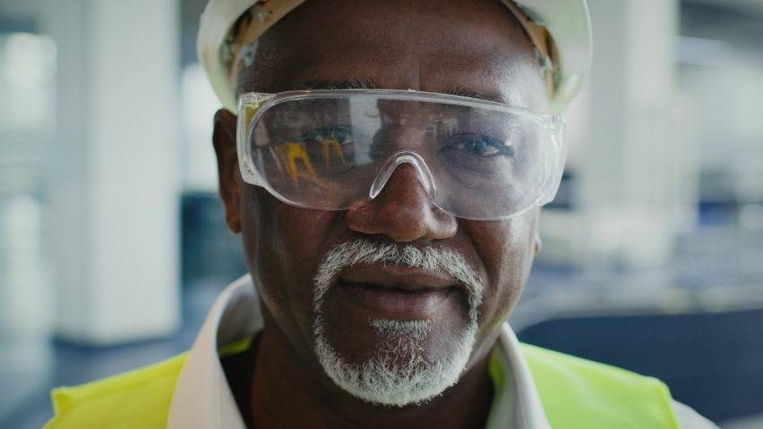 Closeup Portrait Of Mature Black Construction Worker In Protective Helmet, Vest And Eyeglasses Posing Indoors At Site, Smiling African American Civil Engineer Looking At Camera, Slow Motion Footage Royalty-Free Stock Footage #1082735896
