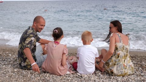 A happy family is sitting on the beach on the seashore in the evening on small stones, they hug together and smile. View from the back.