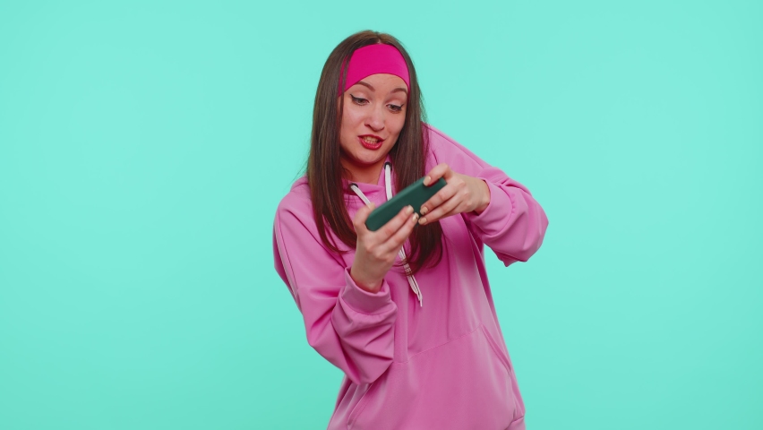 Worried funny addicted woman enthusiastically playing racing video games on mobile phone. Young adult teen girl using smartphone gadget app with drive simulator isolayed on blue studio wall background