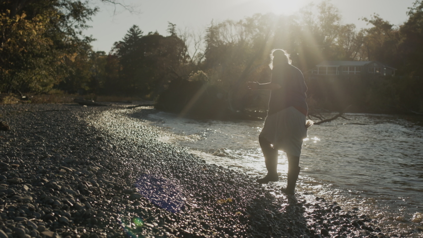 A homeless woman in rubber boots walks along the edge of a lake, looking for something on the shore. Royalty-Free Stock Footage #1082743336