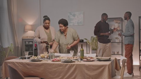 Long shot of two younger and older African American women putting cutlery down on dinner table for holiday celebration, Black men drinking wine and talking on background