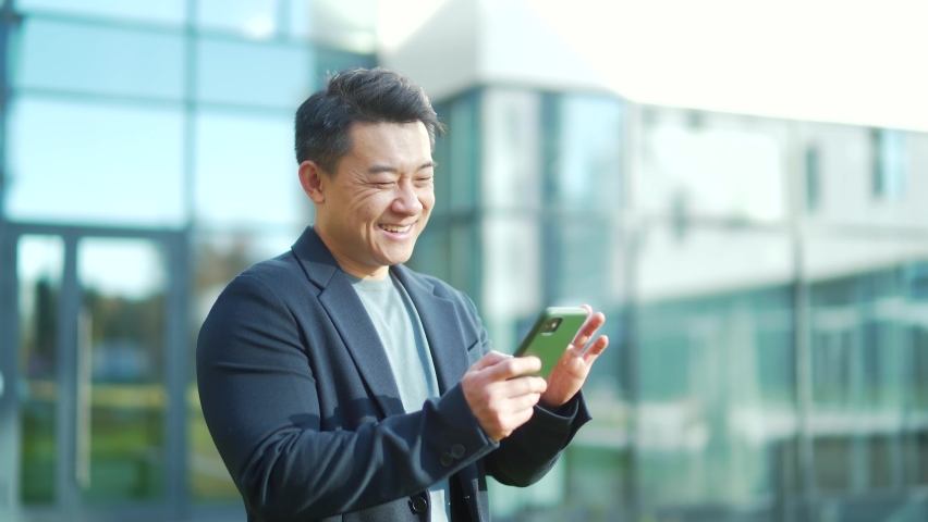 Happy Cheerful asian man enjoys use smartphone standing outside on modern urban building Reads funny news. male smile scrolling, typing, using cellphone. Makes purchases or orders online booking | Shutterstock HD Video #1082755477