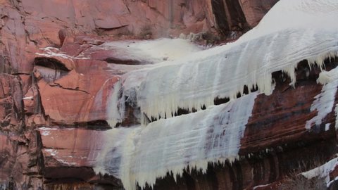 Sheets of ice cover red sandstone ledges that form part of a waterfall in Zion National park during cold winter weather while a small amount of water continues to fall from above. 
