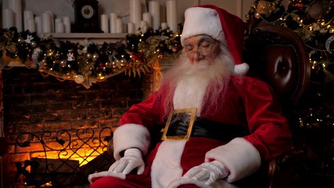 Santa Claus falls asleep while sitting on chair on background fireplace Christmas tree. Old man grandfather dressed as Santa Claus costume sleeping. New Year Christmas winter holidays celebration