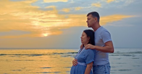 Portrait, a beautiful pregnant woman with a loving husband on the seashore. Man gently hugs a pregnant woman, they smile and are happy. Man and a woman are expecting a baby. Seashore, sunset. 4k