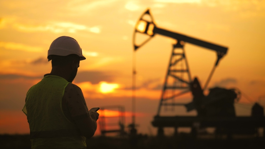 Silhouette working engineer oil rig. Oil rigs at sunset. Silhouette of man engineer with phone overseeing the site of crude oil production at sunset. Industrial, oil and gas concept.