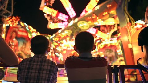Two young brothers next to their mother admiring the spinning carousel at night, at amusement park in France - slow motion
