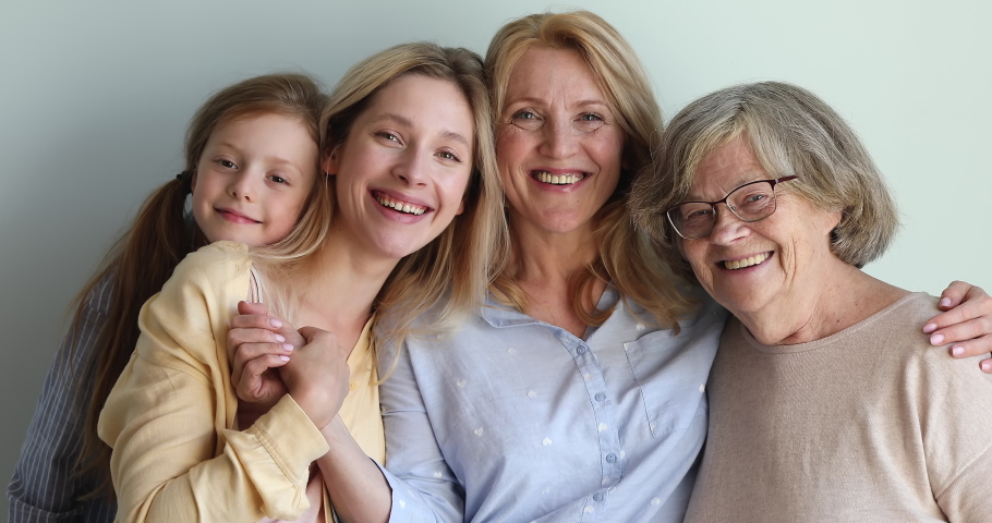 Family of 4 happy pretty females of different generations stand against wall hug look at camera with healthy smiles. Aged woman embrace elderly mom young daughter small granddaughter. Closeup portrait Royalty-Free Stock Footage #1082777059