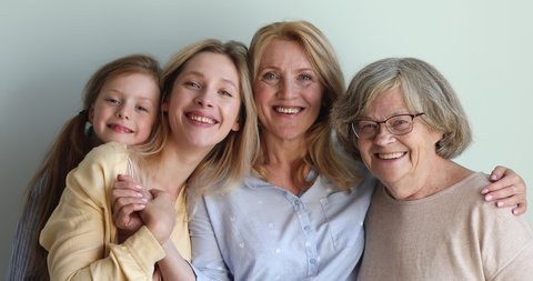 Family of 4 happy pretty females of different generations stand against wall hug look at camera with healthy smiles. Aged woman embrace elderly mom young daughter small granddaughter. Closeup portrait