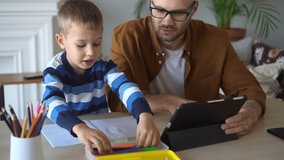 Internet sources of junior children education. Attentive little boy draws with blue marker in copybook while father holds tablet at table close view