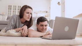 Young couple watching web or video and using device while lying on floor in apartment room. American woman, man look at computer screen and are surprised, talk and lie in light interior. 4k video