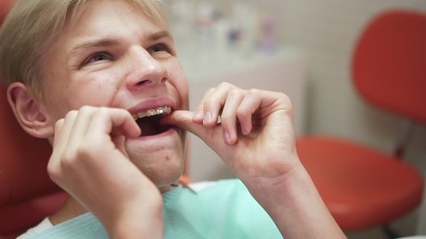 Teenager visits orthodontist. spbd Attractive young man with paper apron tries on aligners trainers sitting in armchair in dental clinic office closeup