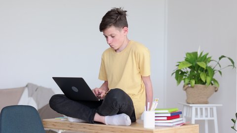 Home education teenage kids. Concentrated teen boy in yellow t-shirt with contemporary laptop sits in lotus pose on wooden table in light room 4k video