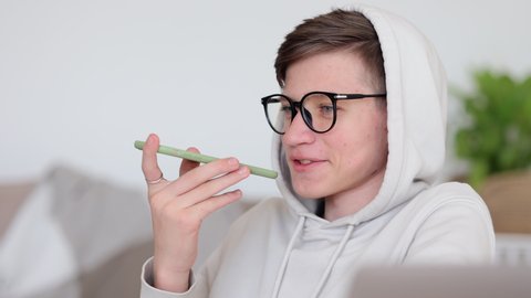 Online education of teen students at home. Schoolboy in white hoodie  with glasses records audio message on smartphone near laptop computer at table closeup 4k video
