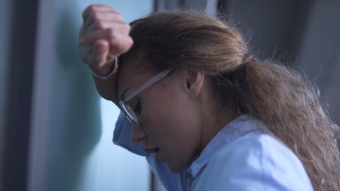 Depressed multiracial woman office worker in eyeglasses banging her head against wall, problems at work, accusation of fraud and forgery, lost important clients and big sum of money, bankruptcy