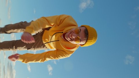 Happy Young man caucasian runs fast amusingly moving his hands with his legs with making funny faces on his face social media wearing yellow hoodie beanie sunglasses close up blue sky summer Lifestyle