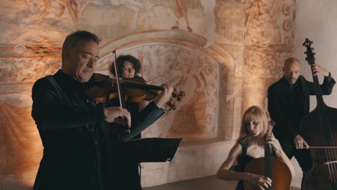 Wide handheld shot of a modern string quartet playing in a medieval style room with a mural in the background
