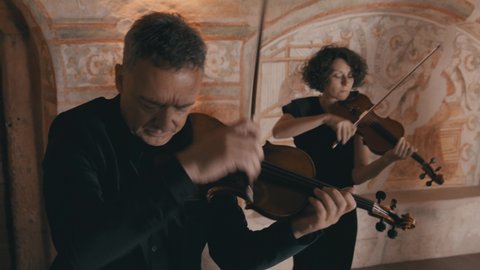 Male violinist plays passionately with a female violinist in the background in a medieval style setting