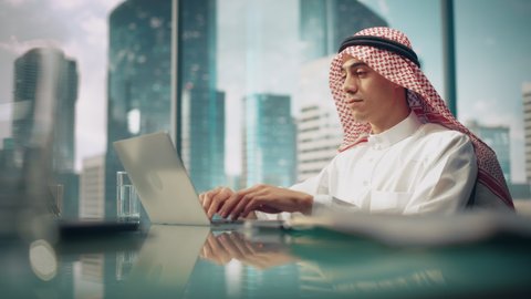 Young Successful Arab Businessman in White Traditional Kandura Sitting in Office and Working on Laptop Computer. Business Manager Planning Investments. Saudi, Emirati, Arab Businessman Concept.